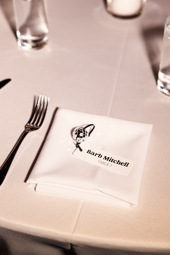 Folded napkin with name card and a small disco ball ornament, beside a fork and water glasses, for wedding reception at River Room