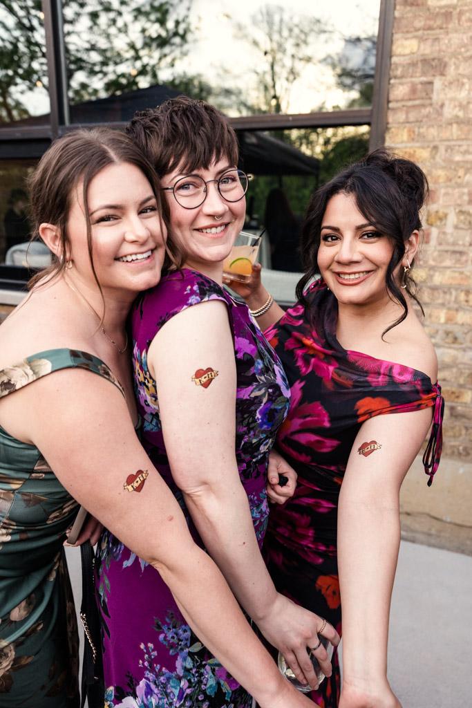Three guests in floral dresses smile, showing matching temporary tattoos on their upper arms outdoors during wedding reception at River Room