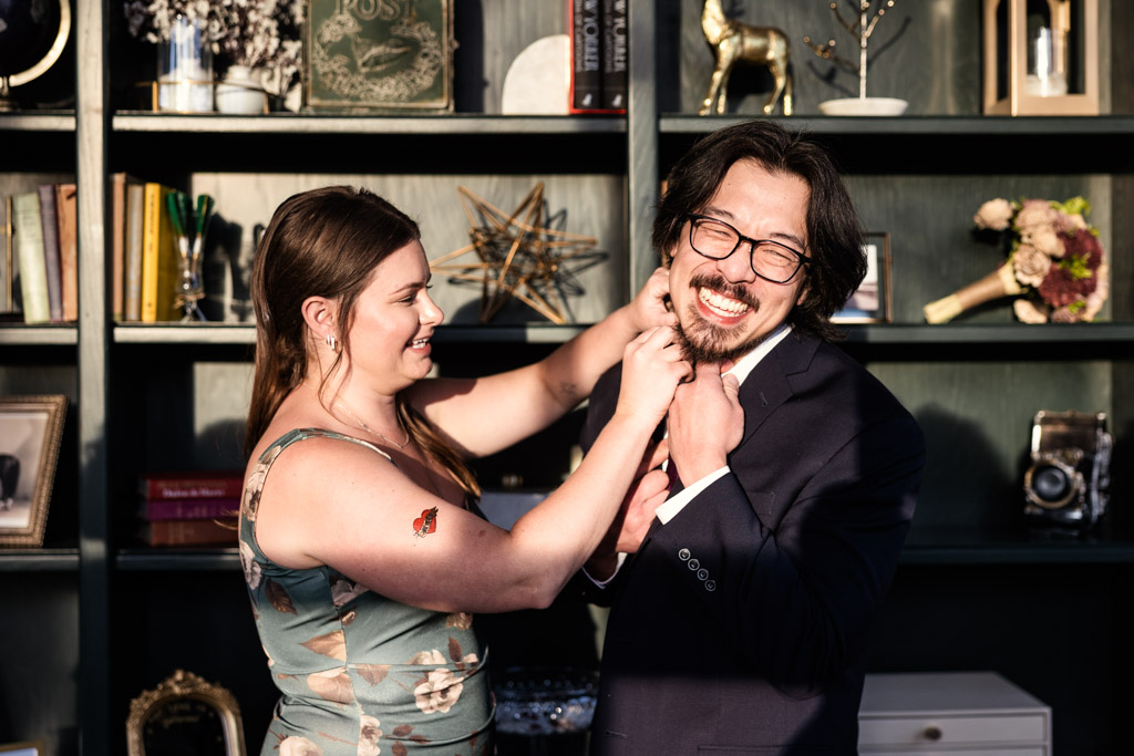 A woman playfully applies a temporary tattoo to a man's neck while both smile, standing in front of a bookshelf at River Room