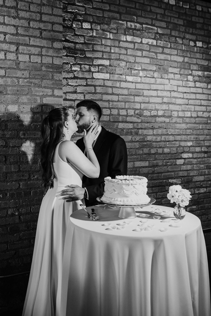 Newlyweds kiss by a table with a cake and flowers, in front of a brick wall inside River Room at Burning Bush Brewery