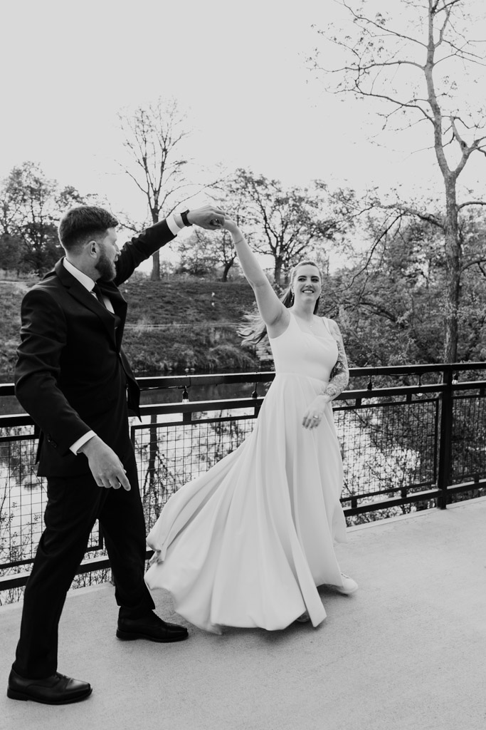 Black and white photo of bride and groom dancing together outdoors on a patio at River Room, with trees and water in the background