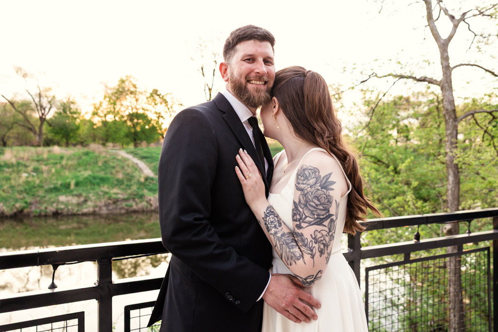 Happy newlyweds in wedding attire embrace on the River Room patio, with lush greenery and a scenic river in the background