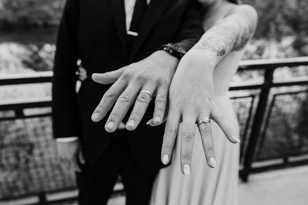 Bride and groom in formal attire show off their wedding rings with hands extended toward the camera