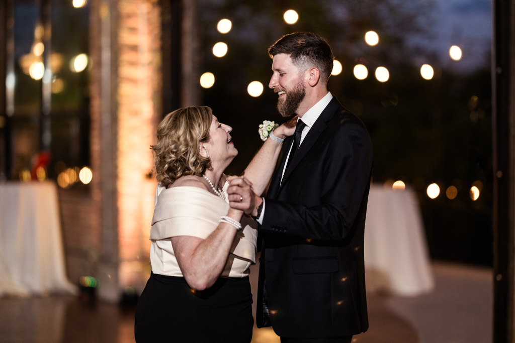 Groom and his mother dancing together at his wedding reception in the River Room, with string lights glowing softly in the background