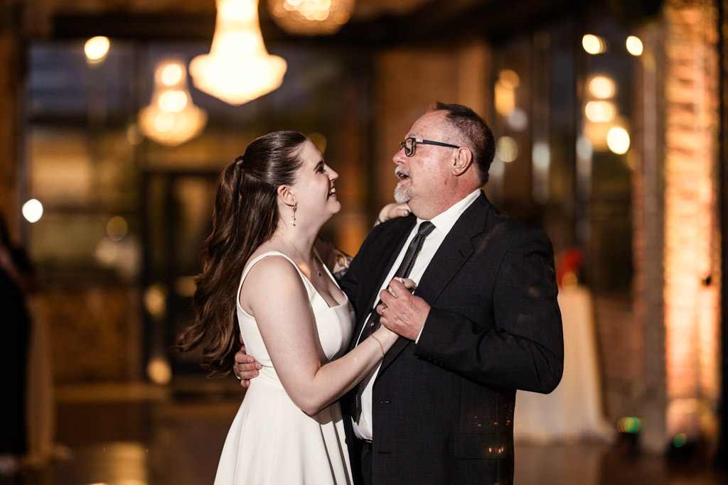 Bride in a white dress dances with her father in a suit at her River Room wedding reception