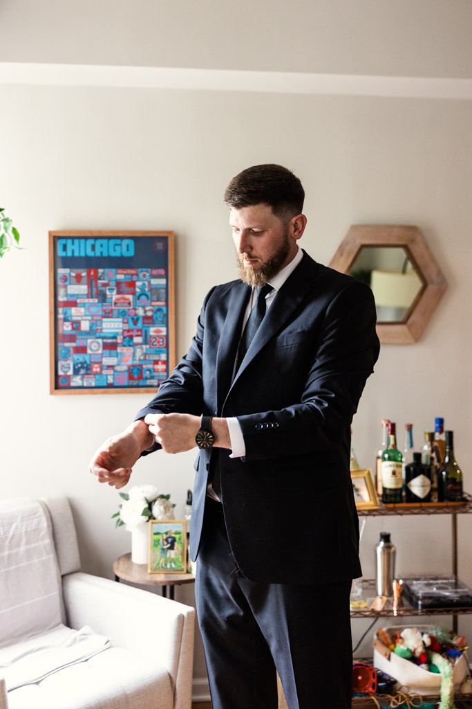 Groom in a suit adjusts cufflinks in his living room, while getting ready for wedding celebration at River Room at Burning Bush Brewery