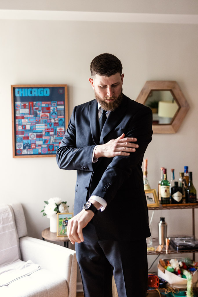 Groom in a dark suit adjusts his sleeve in a living room while getting ready for his wedding at River Room
