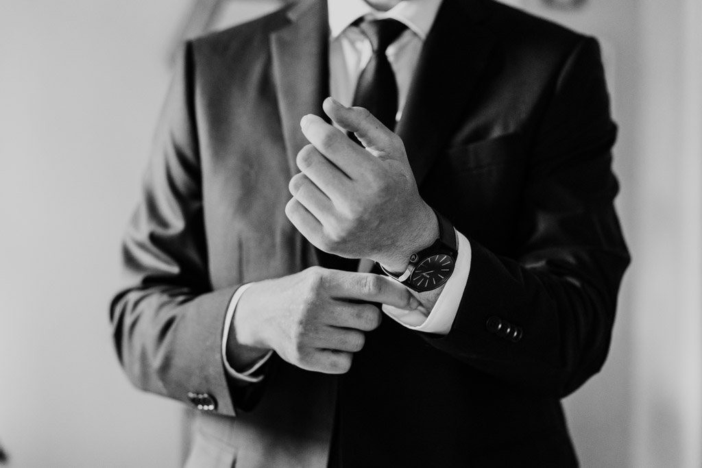Black and white photo of groom in a suit adjusts his watch and cuff with his head partially out of frame