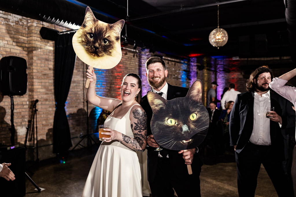 Bride and groom smile holding large cat face cutouts during their wedding celebration at River Room