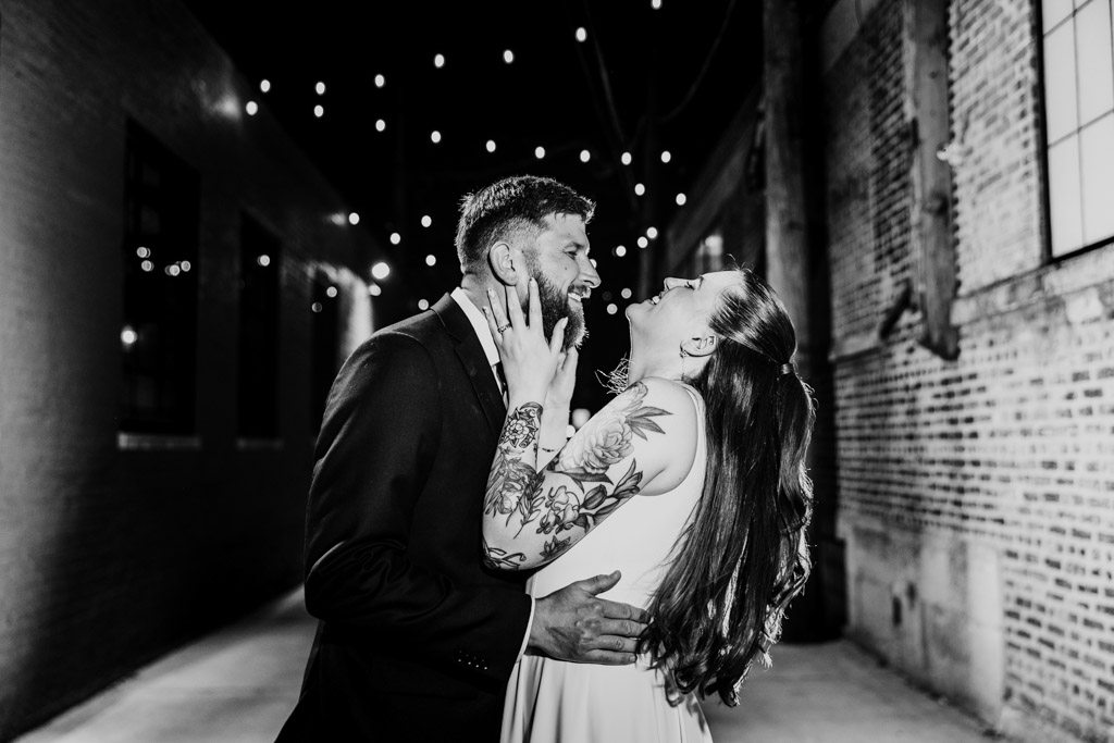 Newlywed couple laughs together at night under string lights in an alley near River Room at Burning Bush Brewery, dressed in formal attire