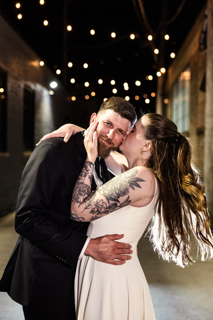 Bride in a white dress kisses groom in a suit under string lights in an alley at night, just steps away from their wedding reception at River Room
