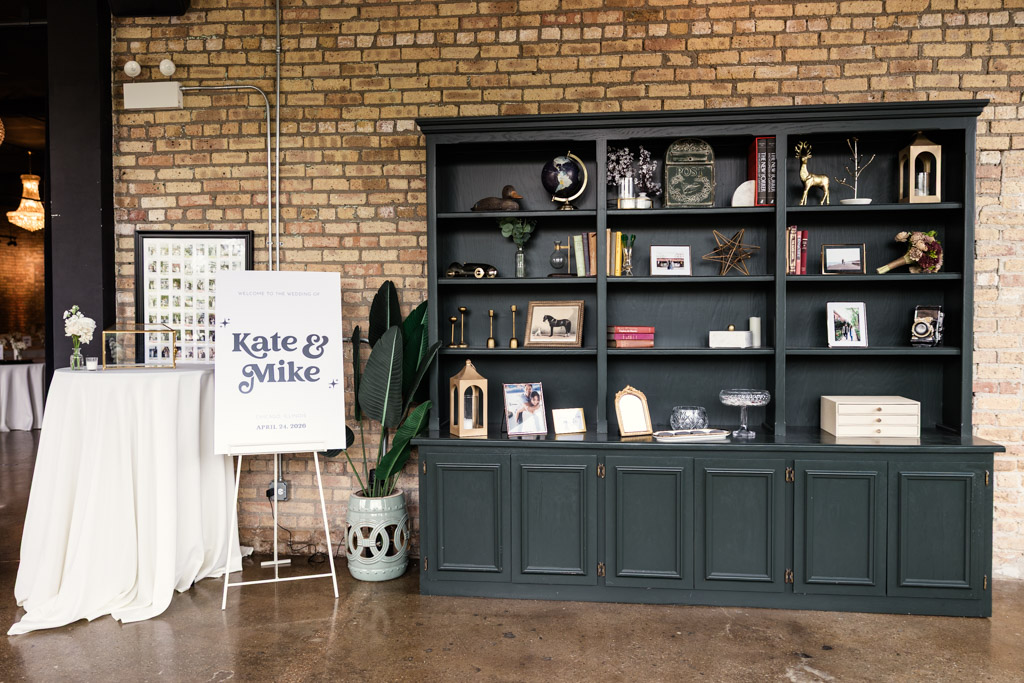 A dark green bookcase with decor stands by a welcome sign for wedding celebration at River Room, set against a brick wall