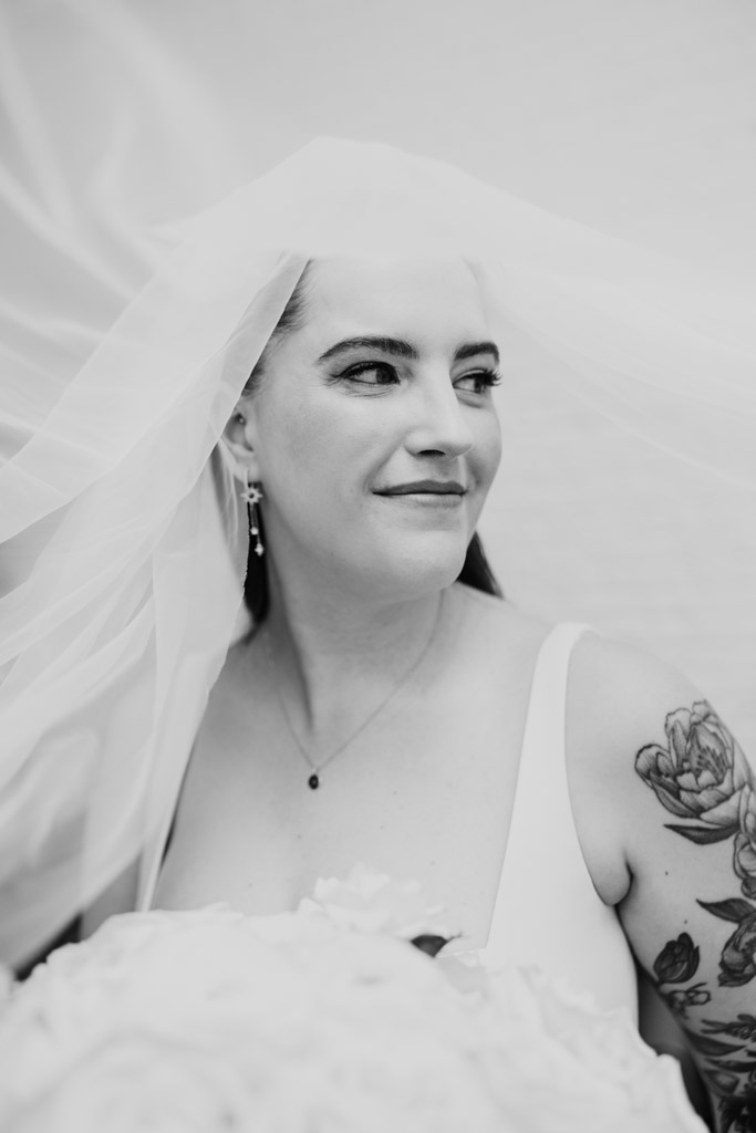 Bride with tattooed arm smiles under her veil, holding a bouquet in a black and white portrait