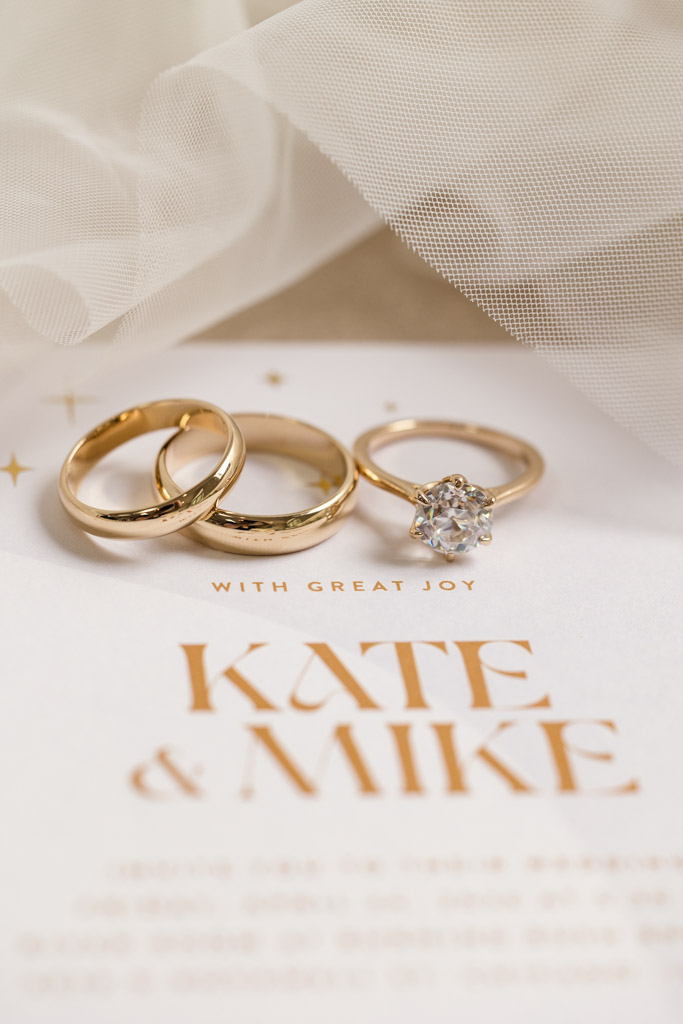 Three gold rings, one with a diamond, rest on a wedding invitation under veil for wedding celebration at the River Room