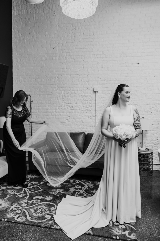Black and white photo of bride holding bouquet smiles as her mother adjusts her long veil