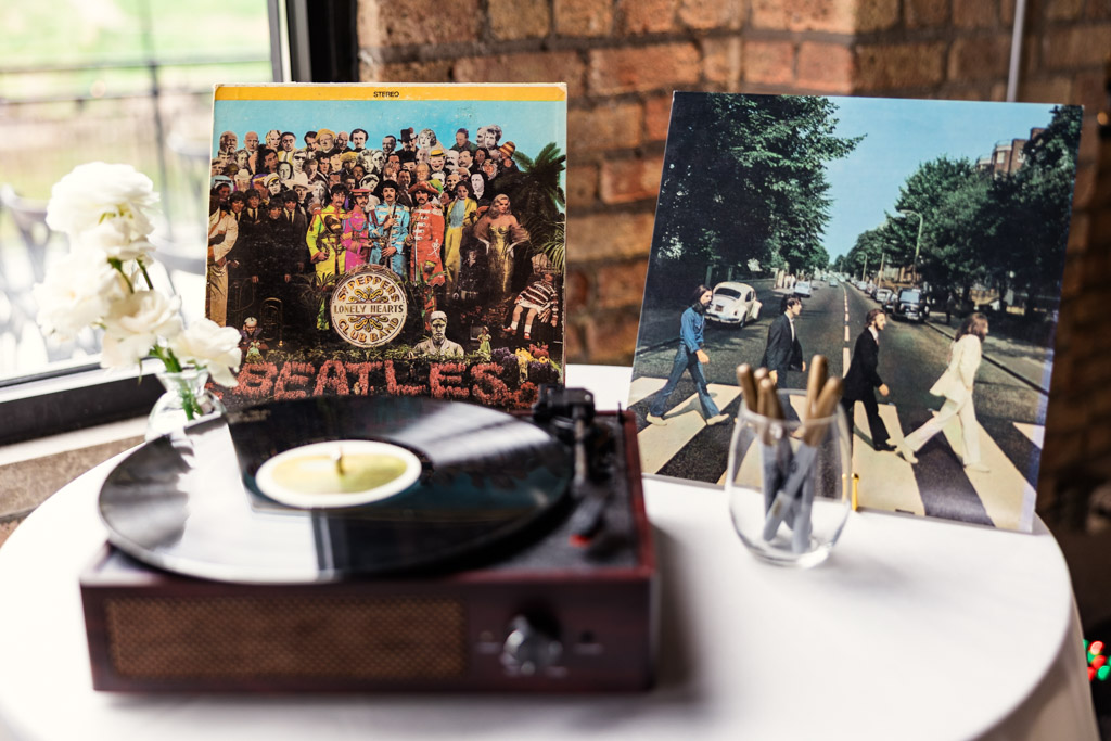 A record player spins a vinyl with Beatles albums displayed near a window accompanied by a vase of white flowers for wedding celebration at RIver Room