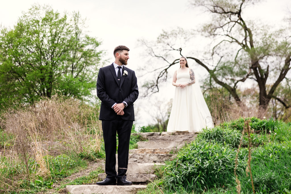 A groom in a suit waits on the stone steps in Horner Park as bride in a white dress approaches from behind him