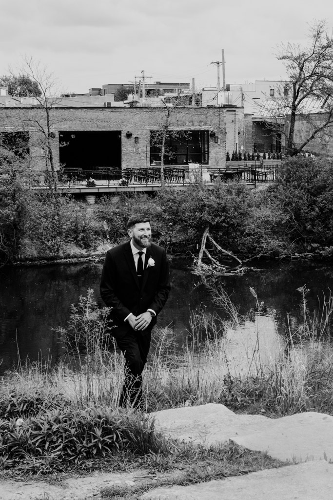 Black and white photo of groom in a suit standing by the riverbank watching the bride walk toward him with buildings and trees in the background