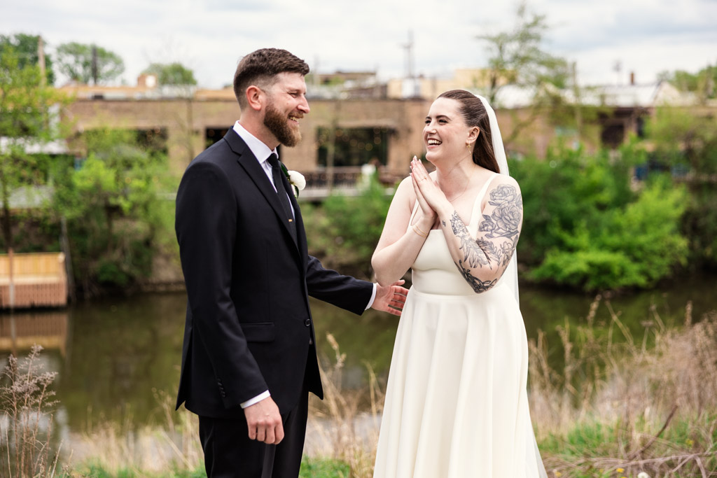 A bride and groom stand outside laughing in Horner Park, surrounded by greenery, just across the river from River Room
