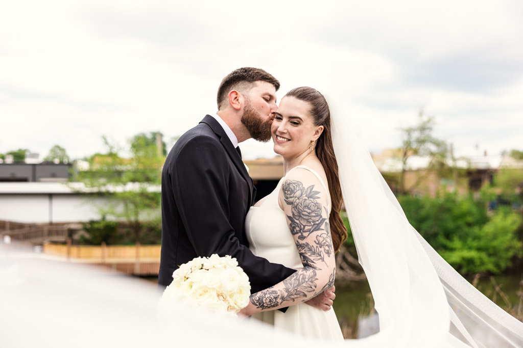Groom kisses bride’s cheek outdoors; bride smiles, holding bouquet, with long veil and floral tattoos near River Room at Burning Bush Brewery