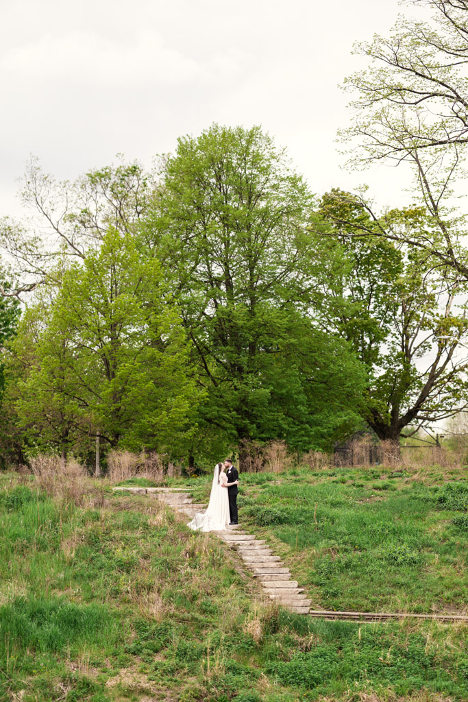 Bride and groom embrace on a wooden path surrounded by green trees and grass during their first look at Horner Park