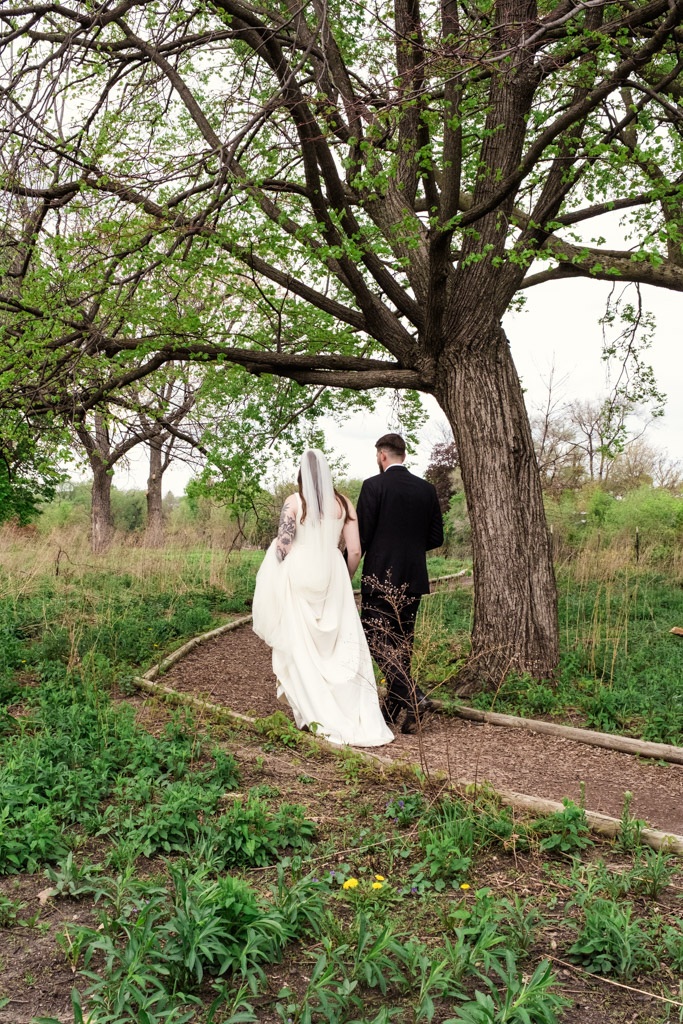 A bride and groom walk together on a dirt path under a large tree in Horner Park