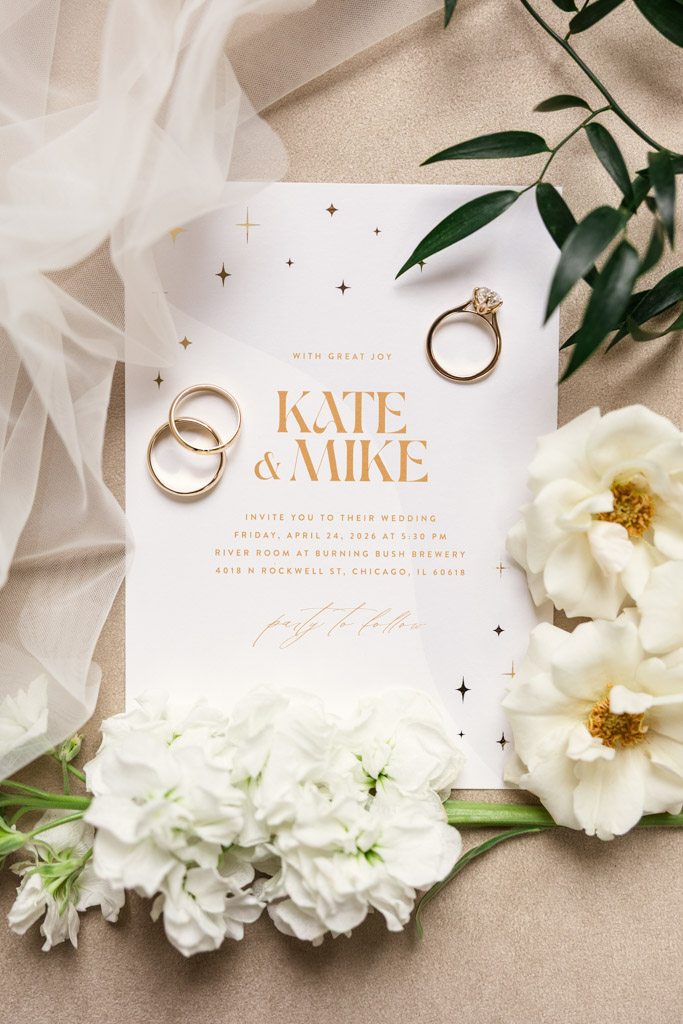 Wedding invitation with white flowers, two rings, and greenery on a white background with gold text for wedding celebration at the River Room