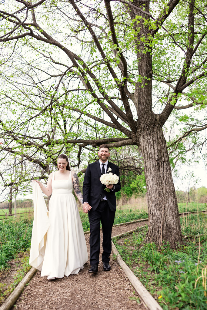 Bride and groom holding hands, walking on a path under leafy trees, bride holding dress and bouquet