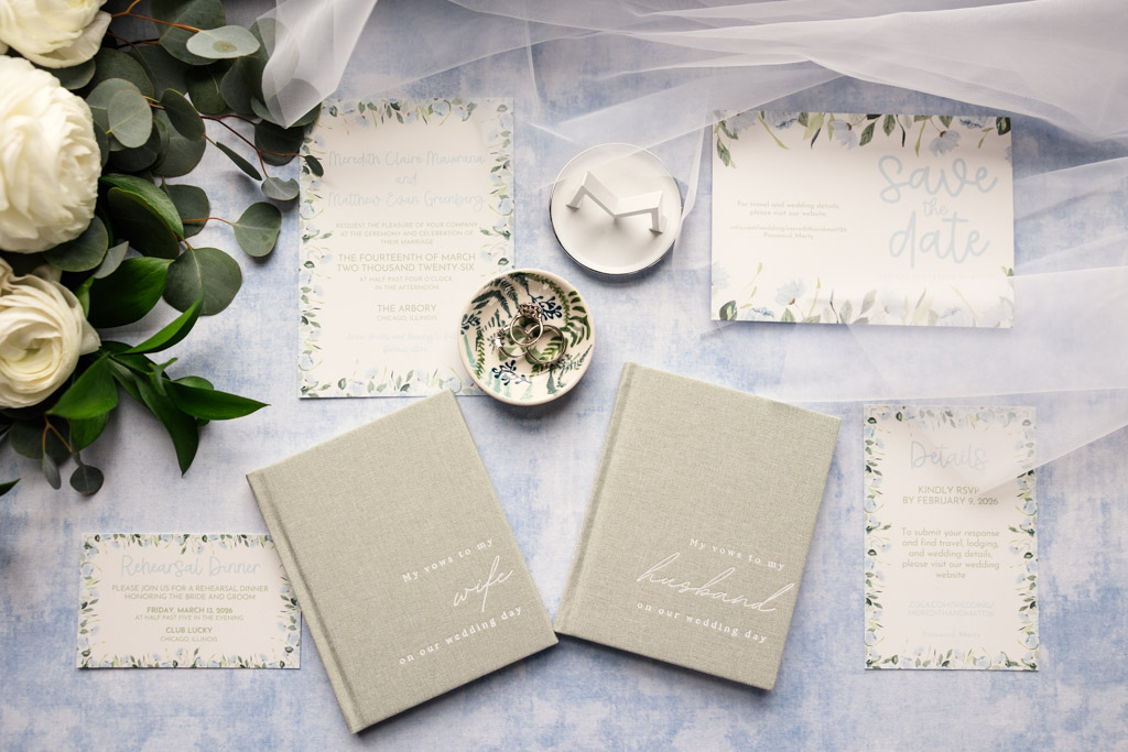 Flat lay of wedding stationery, guest books, rings, and florals on a white cloth with greenery accents for wedding celebration at The Arbory