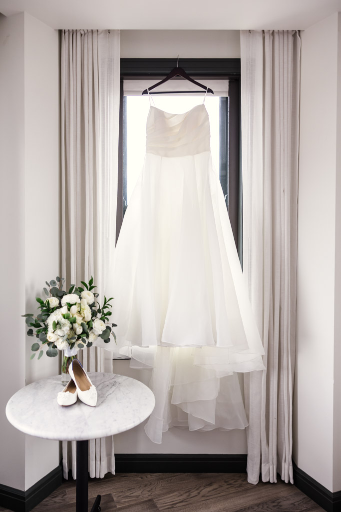 A white wedding dress hangs in a window with bouquet and shoes arranged on a round table below at The Robey Chicago