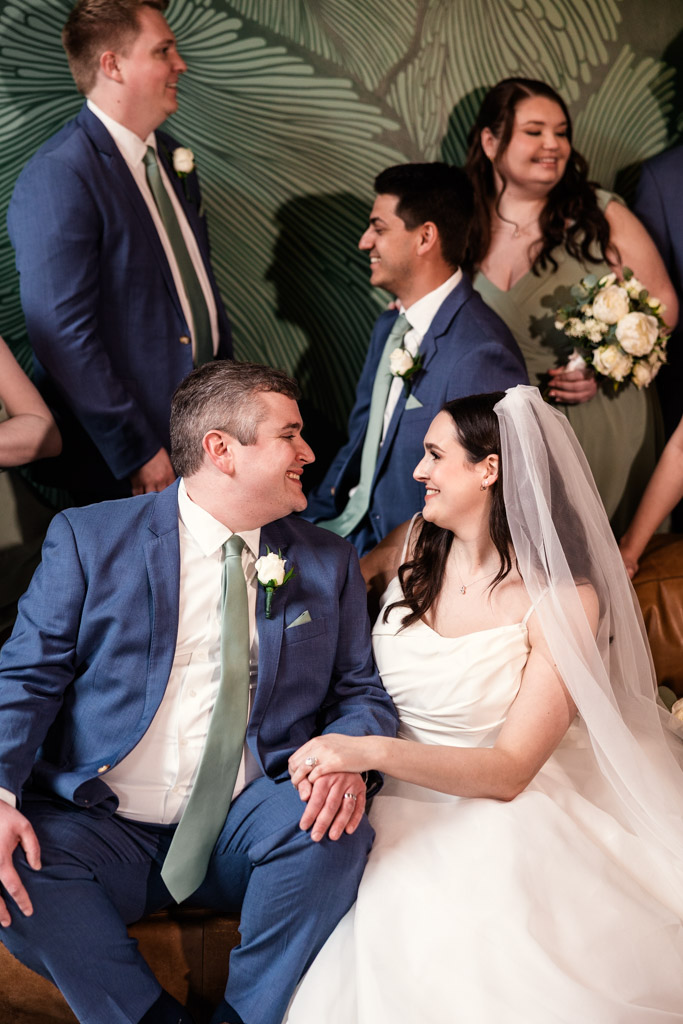 Bride and groom in wedding attire smile at each other at The Arbory, surrounded by bridesmaids in green dresses and groomsmen in blue suits