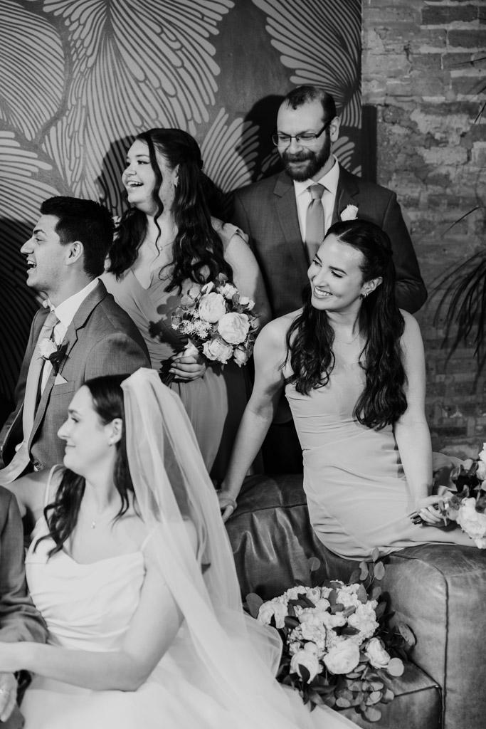 Black and white photo of wedding party members smiling and laughing together indoors at The Arbory, holding bouquets and dressed in formal attire