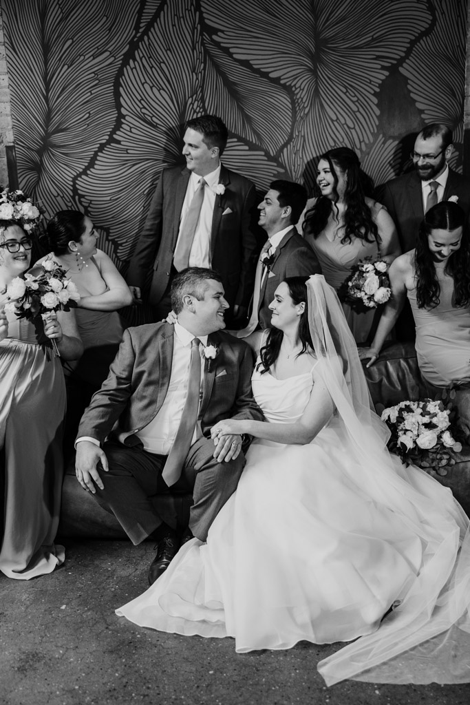 Black and white photo of bride and groom sit surrounded by their wedding party, all smiling and laughing together at The Arbory