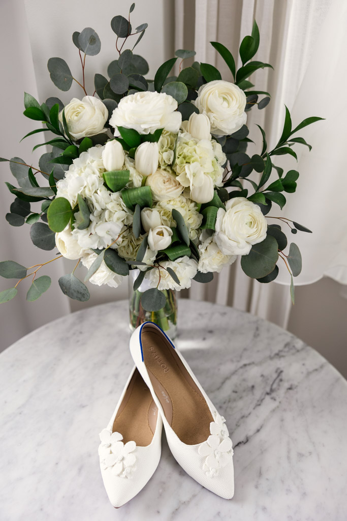 White bridal shoes with floral details rest on a marble table, in front of a lush bouquet of white flowers for wedding celebration at The Robey Chicago