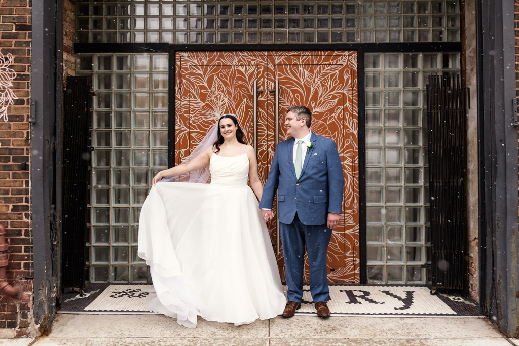 Bride in white dress and groom in blue suit stand smiling in front of the orange floral doors at The Arbory.