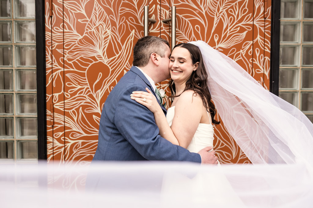 Bride and groom embrace and smile in front of orange floral doors at The Arbory, her veil flowing in the breeze