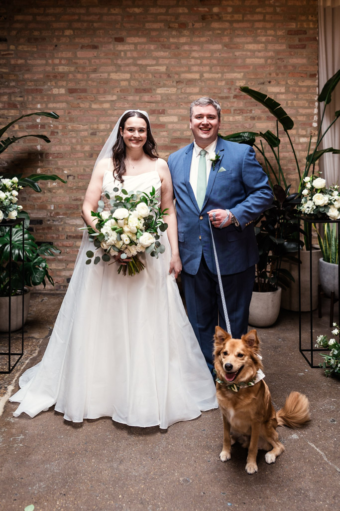 Portrait of bride and groom with their dog on a leash at The Arbory, surrounded by white flowers and green plants