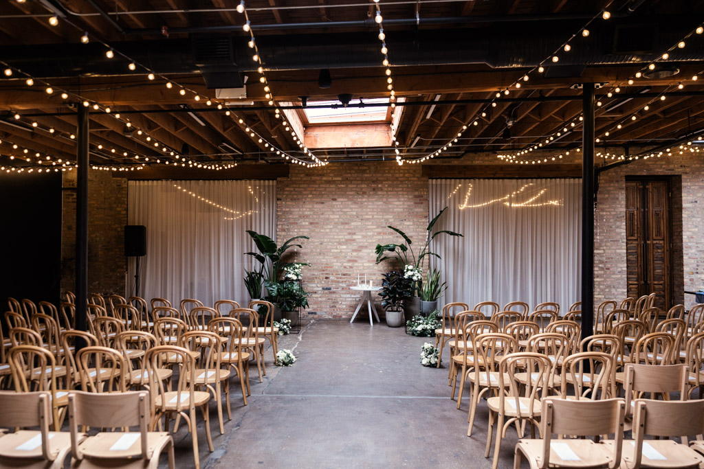 Wedding ceremony space at The Arbory, features string lights, wooden chairs, white drapery, and lush greenery