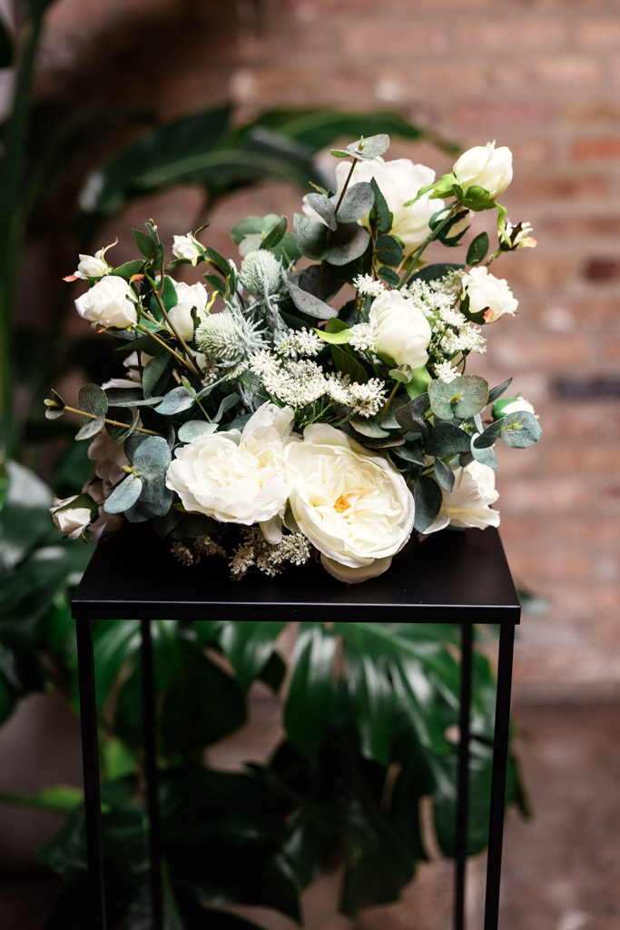 White roses and greenery in a floral arrangement on a black stand for wedding celebration at The Arbory, with a brick wall in the background