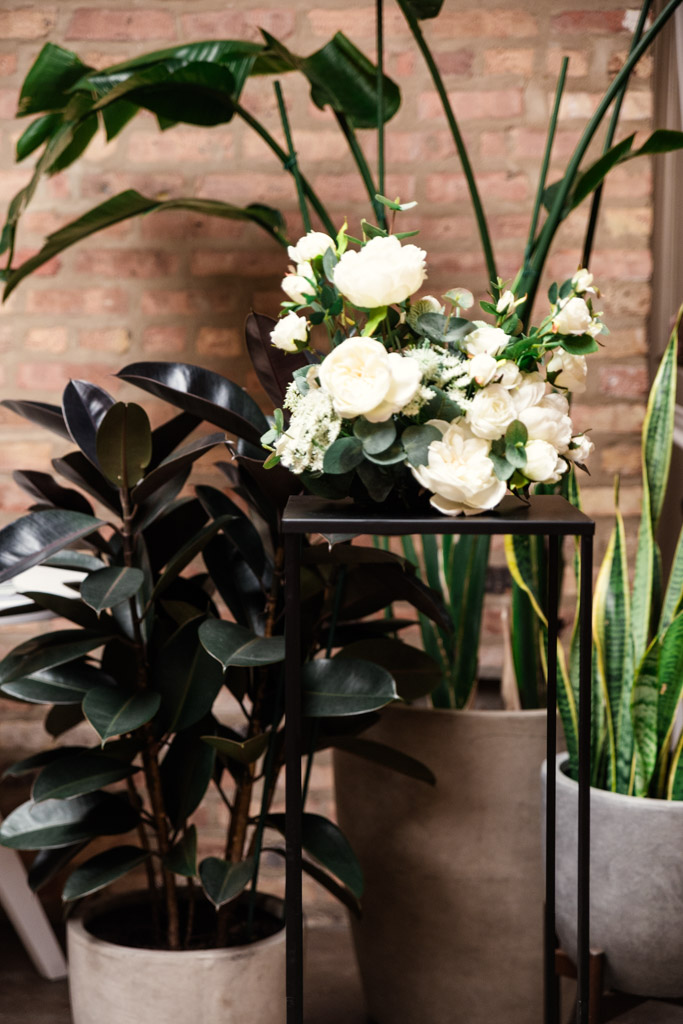 A bouquet of white flowers on a black stand, surrounded by potted green plants against a brick wall for wedding celebration at The Arbory