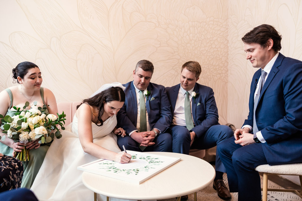 A bride signs a ketubah at a table at The Arbory, surrounded by the groom, her bridesmaid and two groomsmen in blue suits