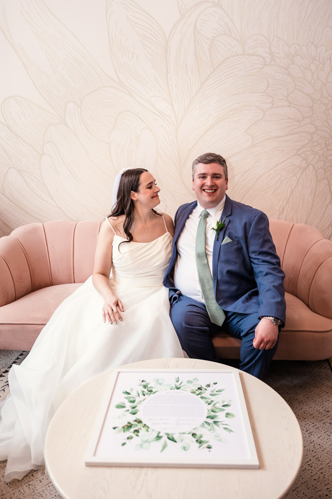 A bride and groom sit smiling on a pink sofa at The Arbory, with their ketubah on the table in front of them
