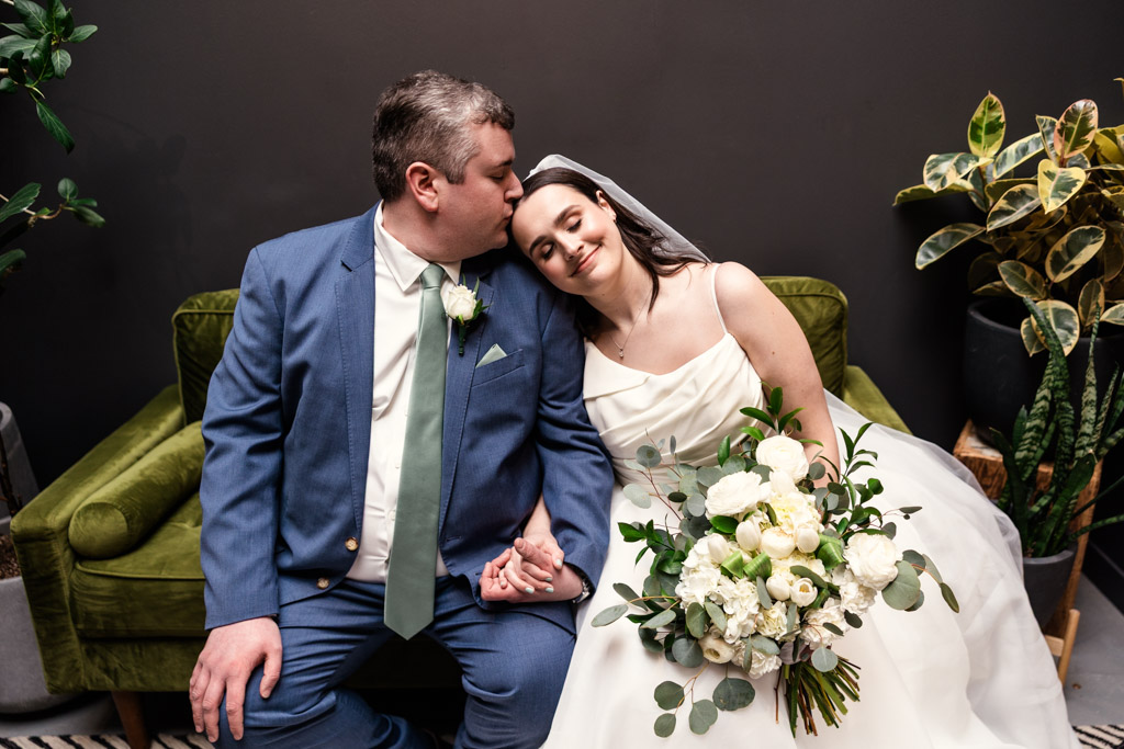 Groom kisses bride's head as they sit on a green couch, holding hands and smiling, bride holding a bouquet of white flowers