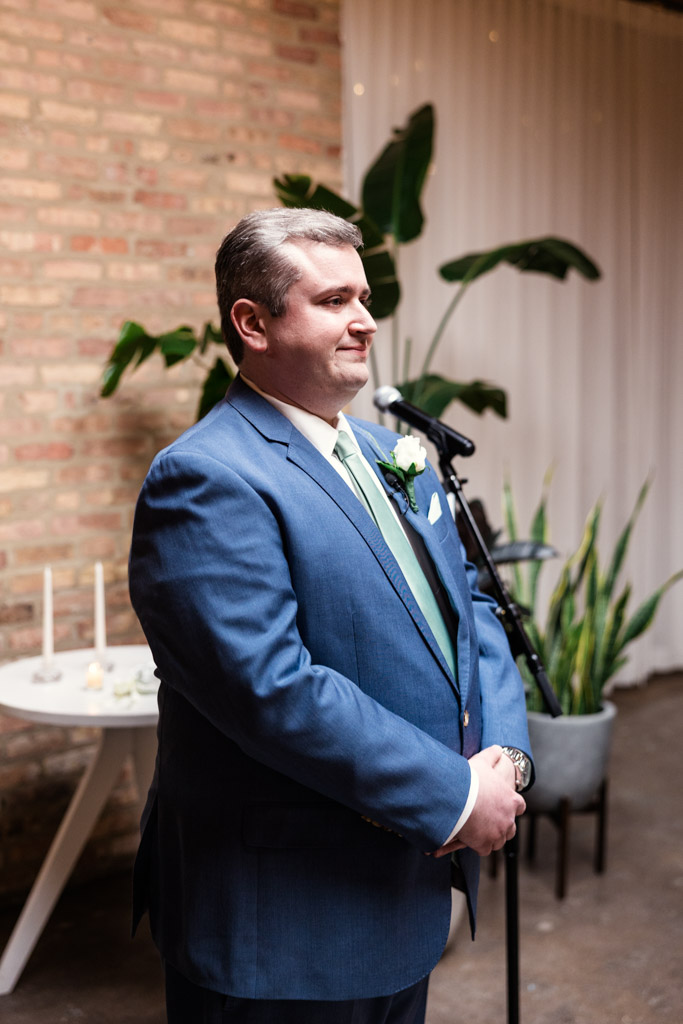 Groom in blue suit stands at the front of his wedding ceremony at The Arbory, with plants and a brick wall in the background