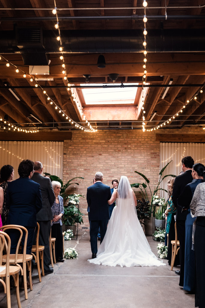Bride walks down the aisle with her father in a warmly lit indoor wedding ceremony at The Arbory
