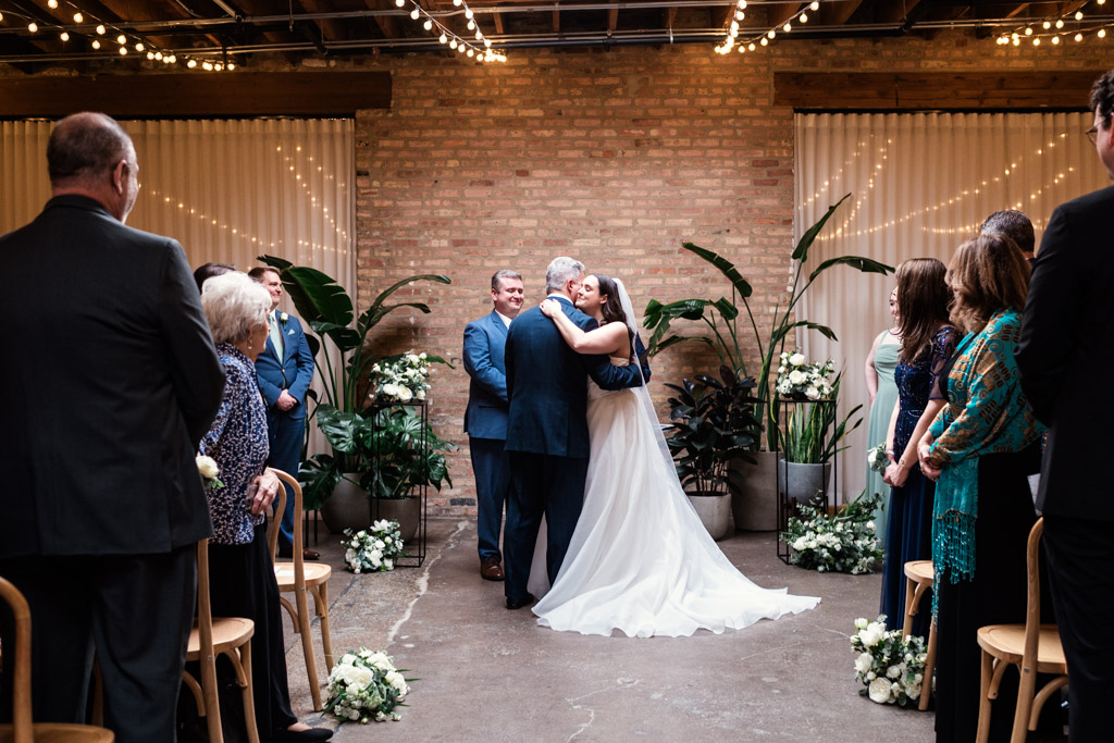 Bride and her father hug at indoor wedding ceremony with guests watching, surrounded by flowers and string lights at The Arbory
