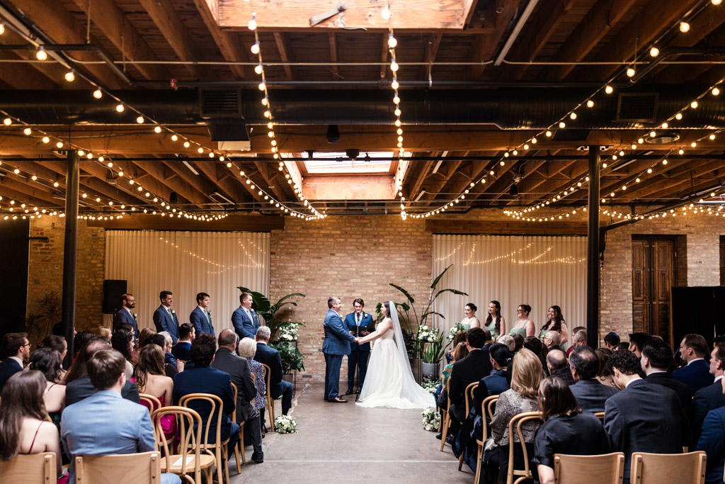 Bride and groom hold hands under string lights during their wedding ceremony at The Arbory, surrounded by their wedding party and seated guests