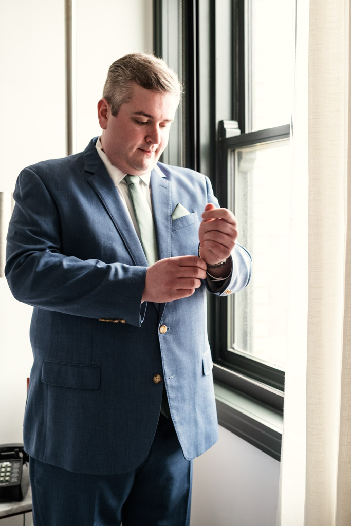 Man in a blue suit adjusts his cuff while standing by a window with natural light at The Robey Chicago