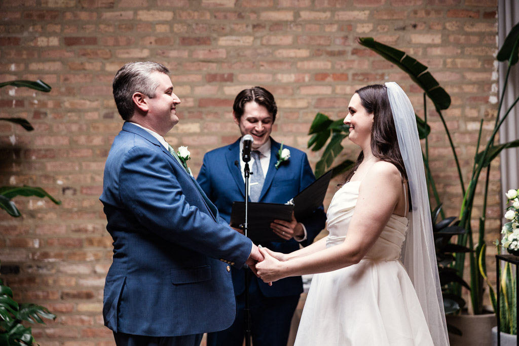 Bride and groom hold hands, smiling at each other, during their wedding ceremony with an officiant behind them at The Arbory