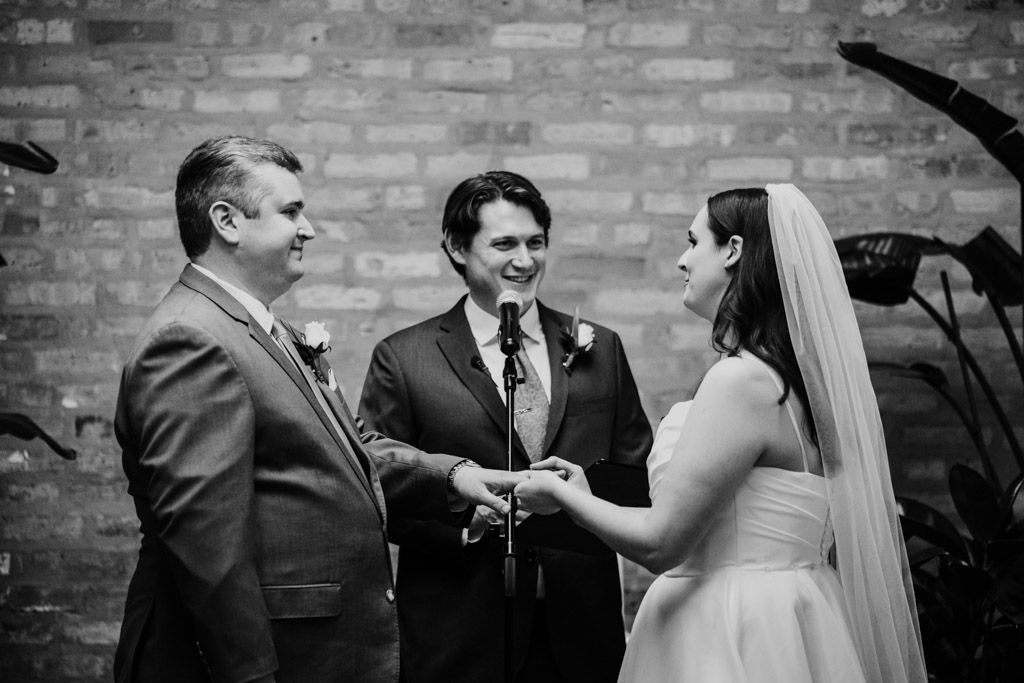 Black and white photo of bride placing ring on groom's finger during their wedding ceremony at The Arbory, officiant smiling between them